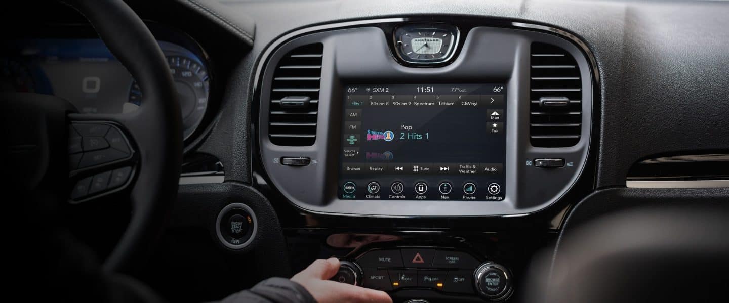 Primer plano de la pantalla táctil del Chrysler 300 2023 con la mano de un conductor alcanzando los controles que hay debajo de ella.