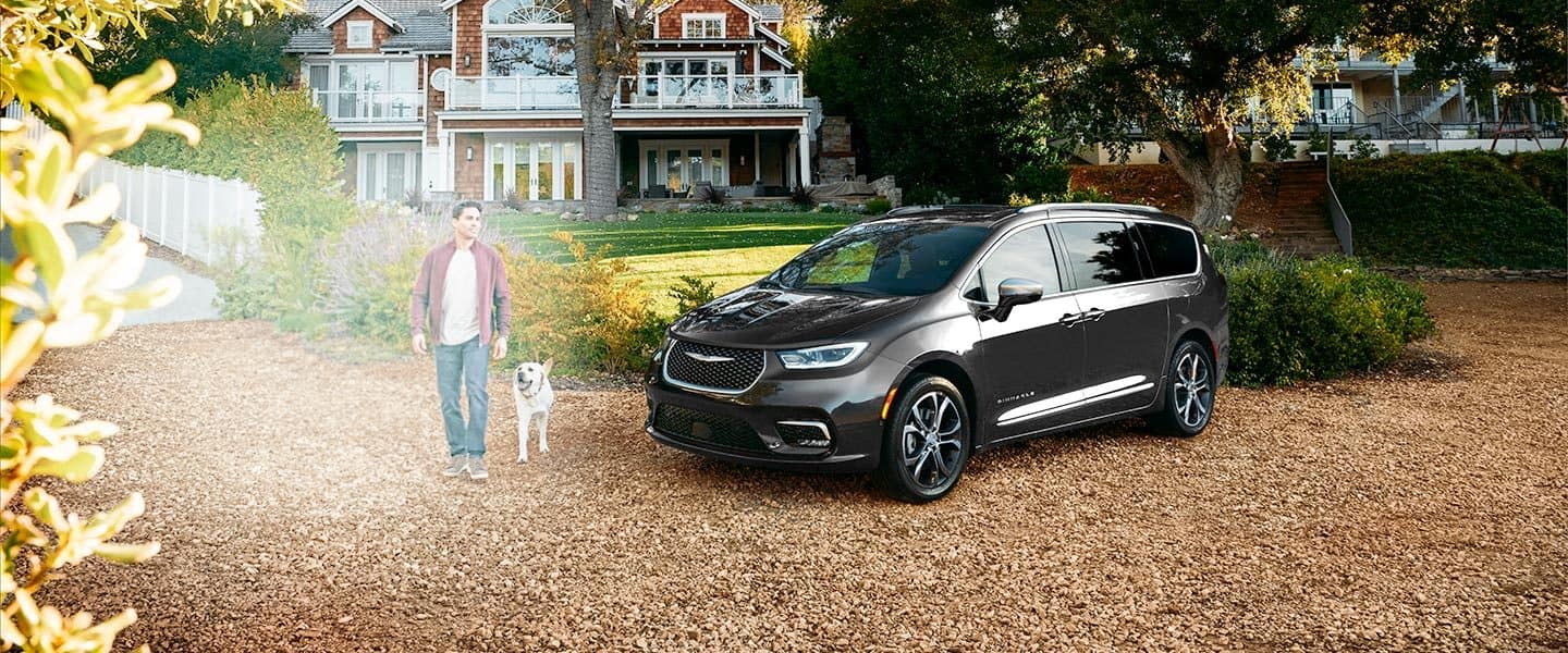 Una Chrysler Pacifica Pinnacle 2023 estacionada en la entrada de garaje de un hogar palaciego, con un hombre y un perro caminando cerca.