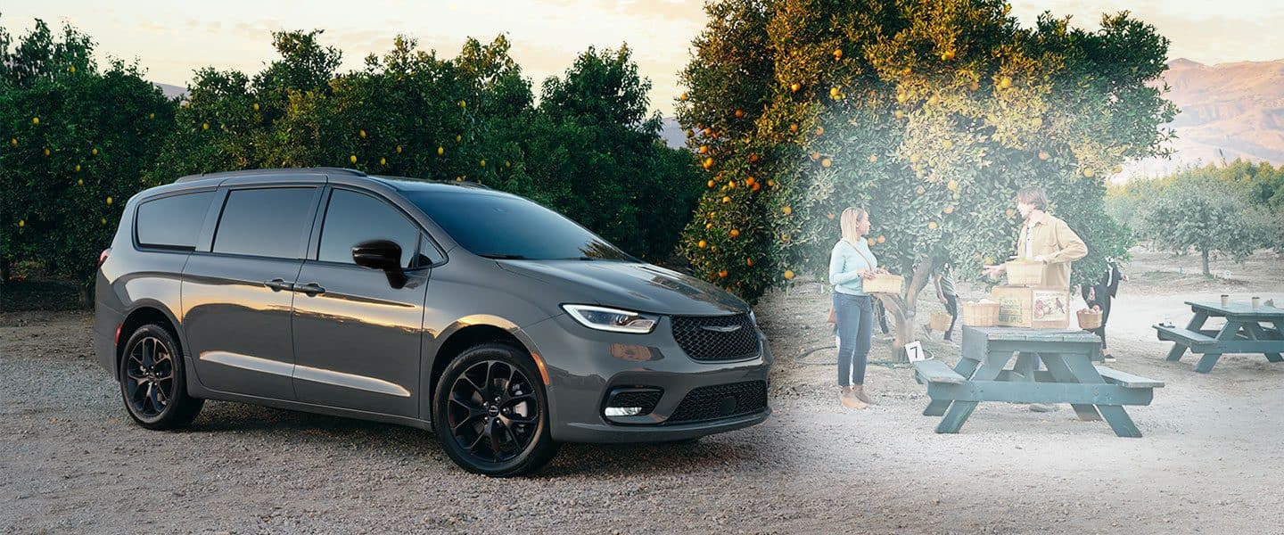 Vista de perfil en ángulo de la Chrysler Pacifica Limited 2023, con el paquete de apariencia S disponible, estacionada en una arboleda naranja al atardecer, con un hombre y una mujer parados cerca.