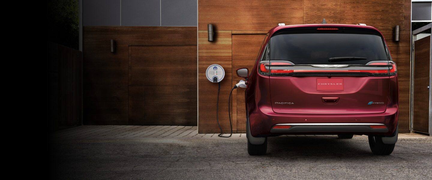 Vista trasera de la Chrysler Pacifica TBD Hybrid 2023 estacionada fuera de un garaje, con un cable de carga enchufado en su puerto de carga.