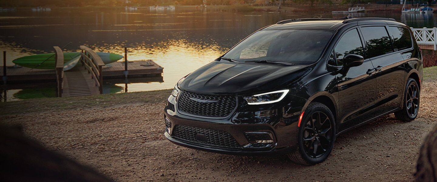 Un perfil en ángulo del lado del conductor de una Chrysler Pacifica Limited 2023 con el paquete de apariencia S estacionado en la costa, junto a un muelle al atardecer.