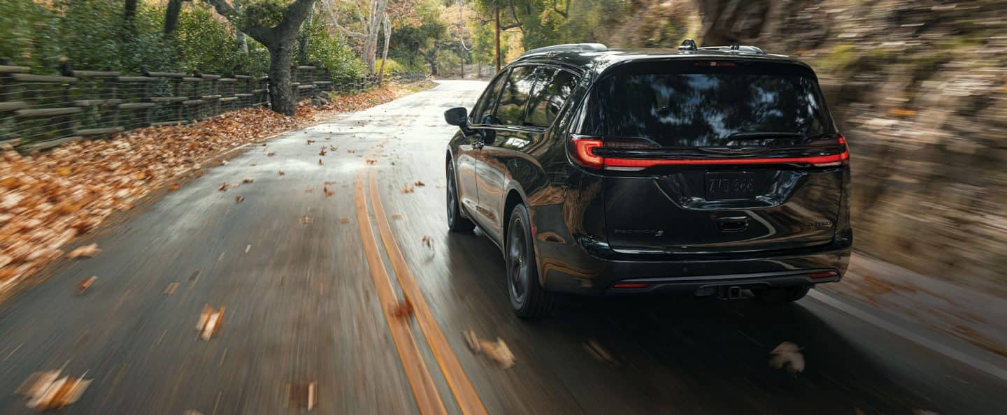 A rear angle of a 2023 Chrysler Pacifica Limited with the S Appearance Package being driven on a country road. The background is blurry indicating the vehicle is in motion. 