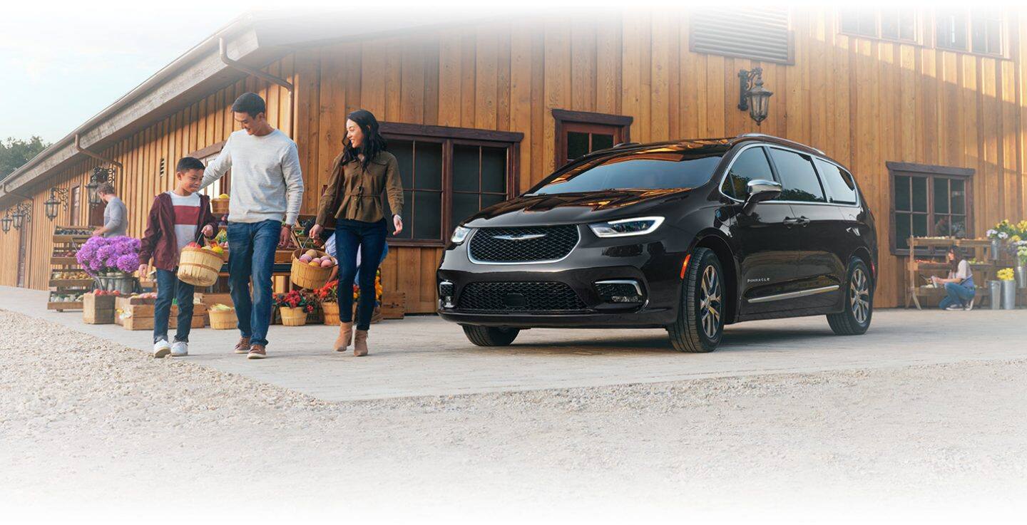 Una Chrysler Pacifica Pinnacle Plug-In Hybrid 2024 negra estacionada junto a un mercado de agricultores con una familia de tres personas que camina cerca llevando cestas de productos.