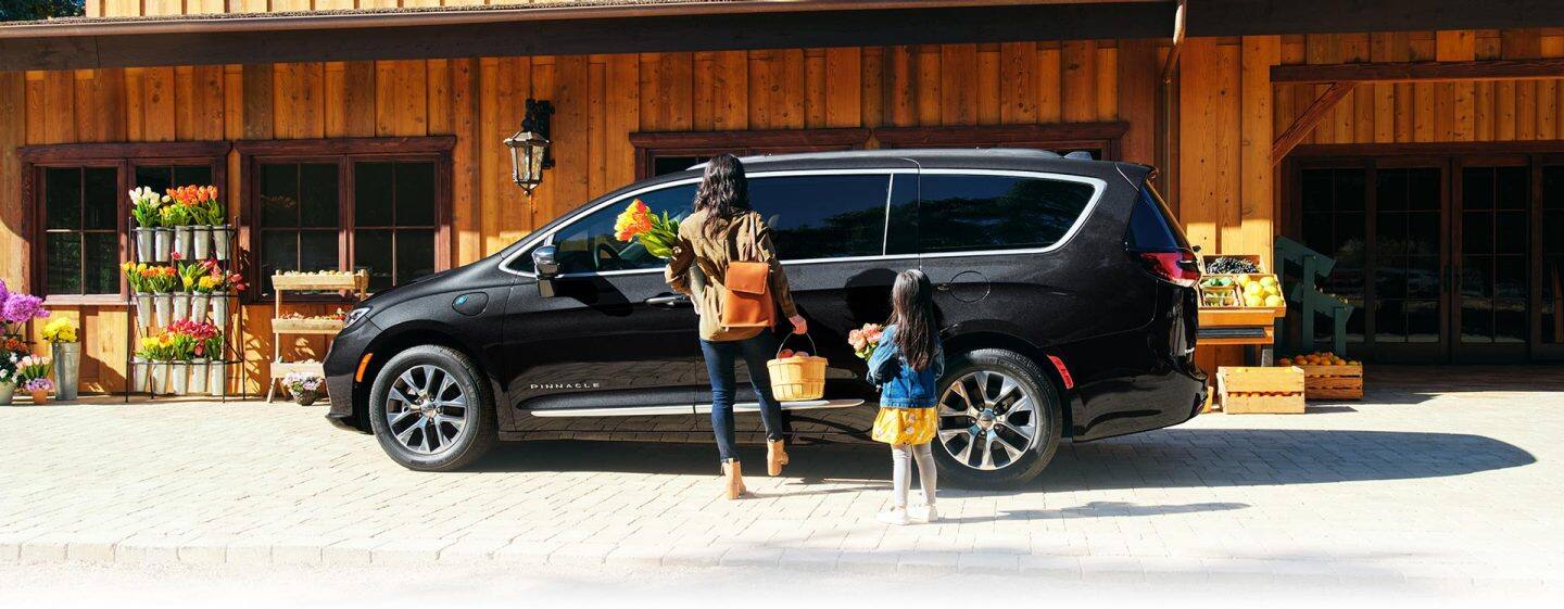 Madre e hija con las manos llenas y de pie junto a la puerta lateral corrediza eléctrica manos libres de una Chrysler Pacifica Pinnacle Hybrid 2023 negra estacionada en un mercado de agricultores, mientras la madre desliza el pie por debajo de la puerta. Logo de Evento de ventas de Fin de año.