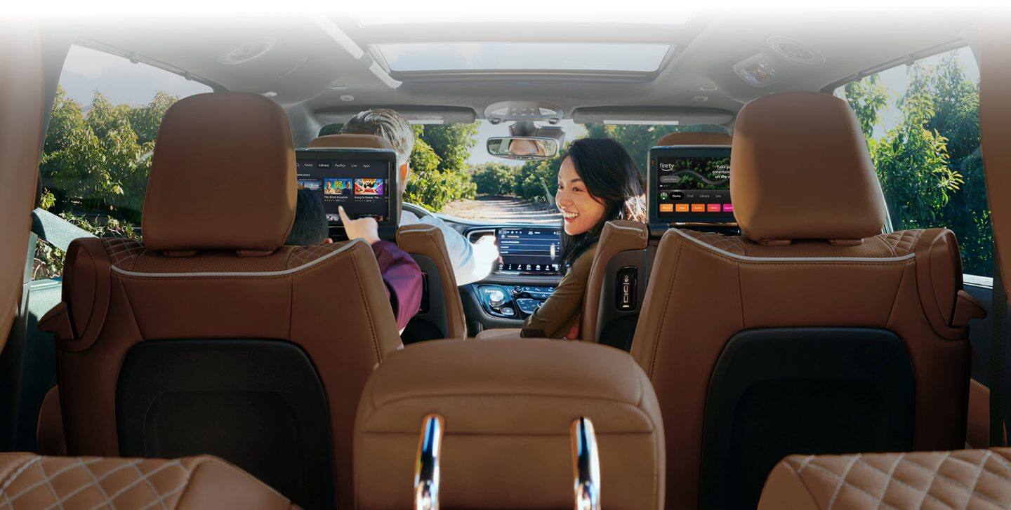 La pasajera delantera de la Chrysler Pacifica Pinnacle Hybrid enchufable 2024 se da vuelta para mirar a un niño en la segunda fila, que explora opciones en la pantalla táctil colocada en el respaldo del asiento delantero.