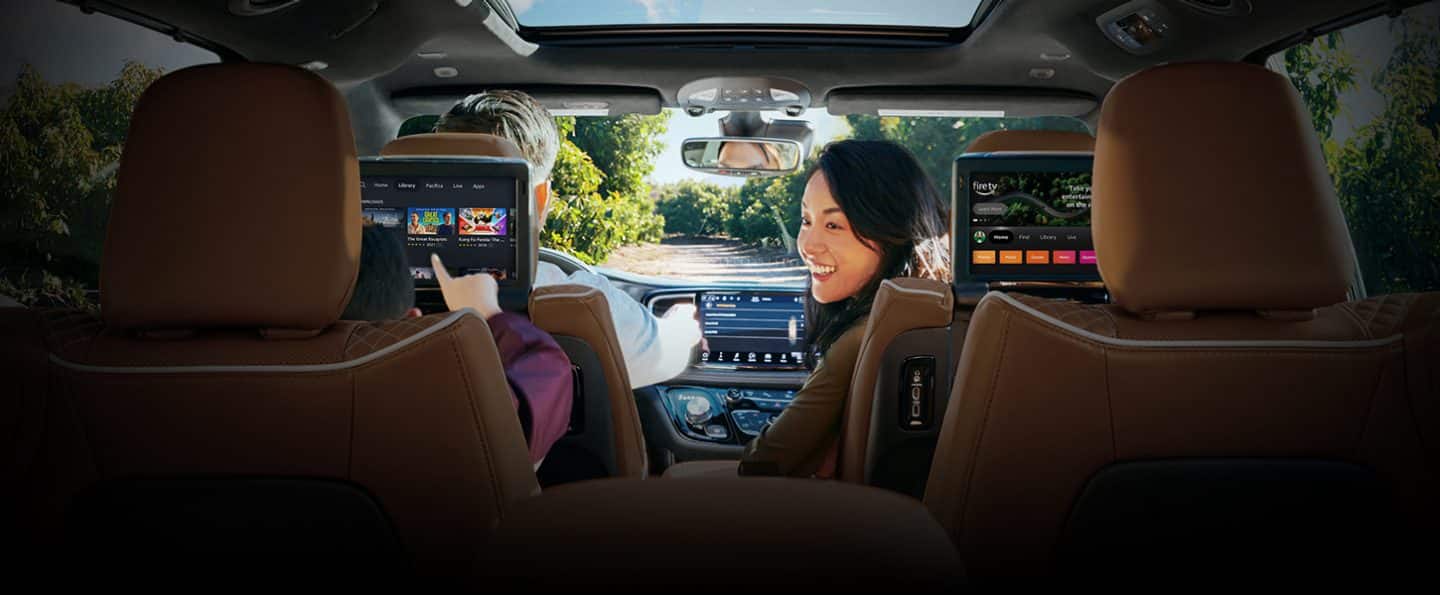 The front passenger in the 2024 Chrysler Pacifica Pinnacle turning to look at a child in the second row who is browsing options on the front seatback-mounted touchscreen.