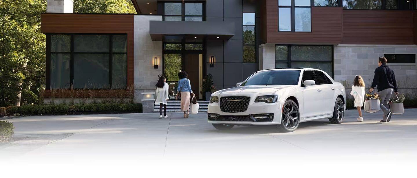 Un Chrysler 300 300S blanco 2023 estacionado en el camino de entrada de una amplia casa contemporánea, con una familia de cuatro personas caminando desde el vehículo hacia la puerta principal.