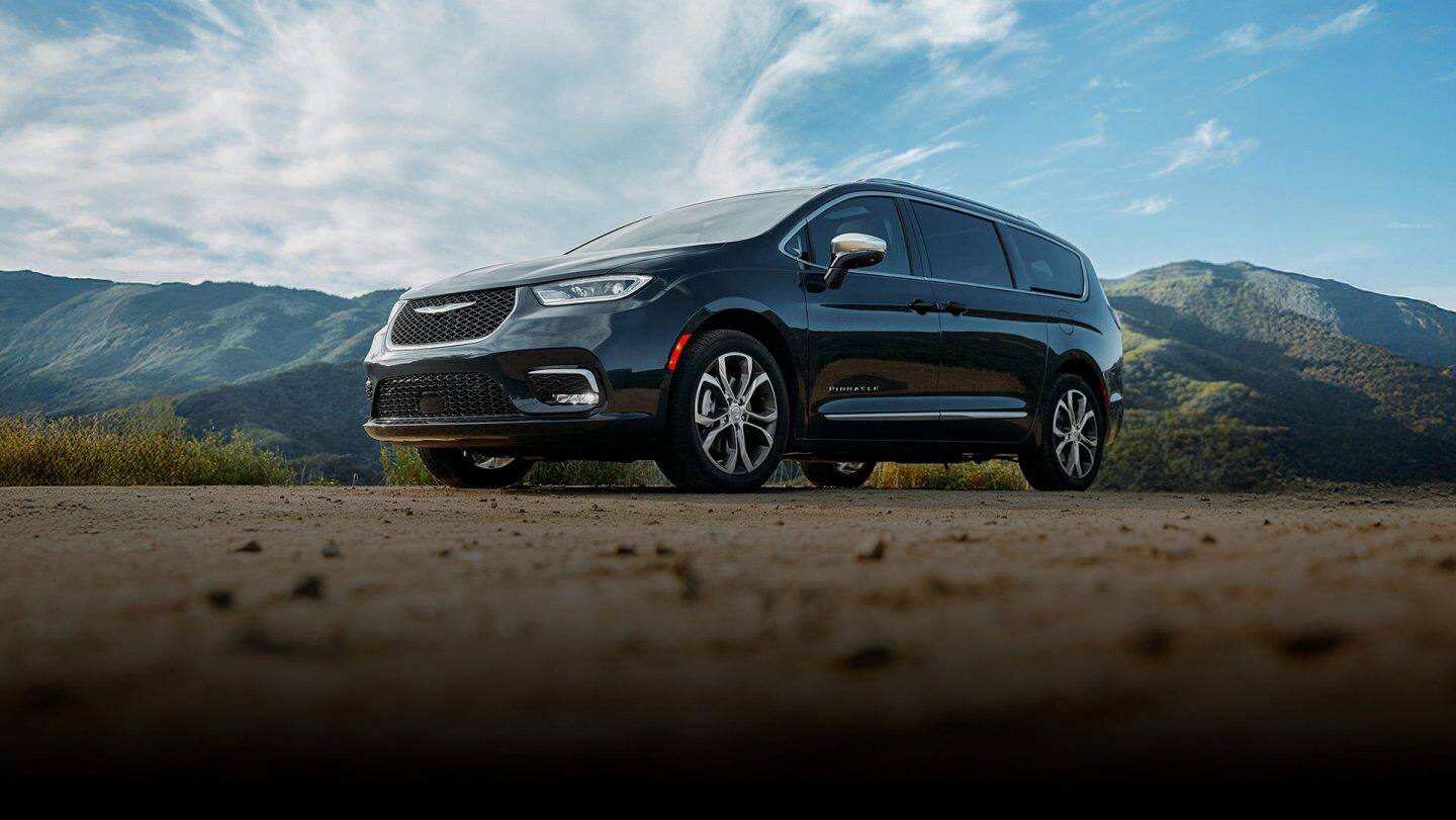 A driver-side front angle of a black 2026 Chrysler Pacifica Pinnacle parked on a clearing with mountains in the background.