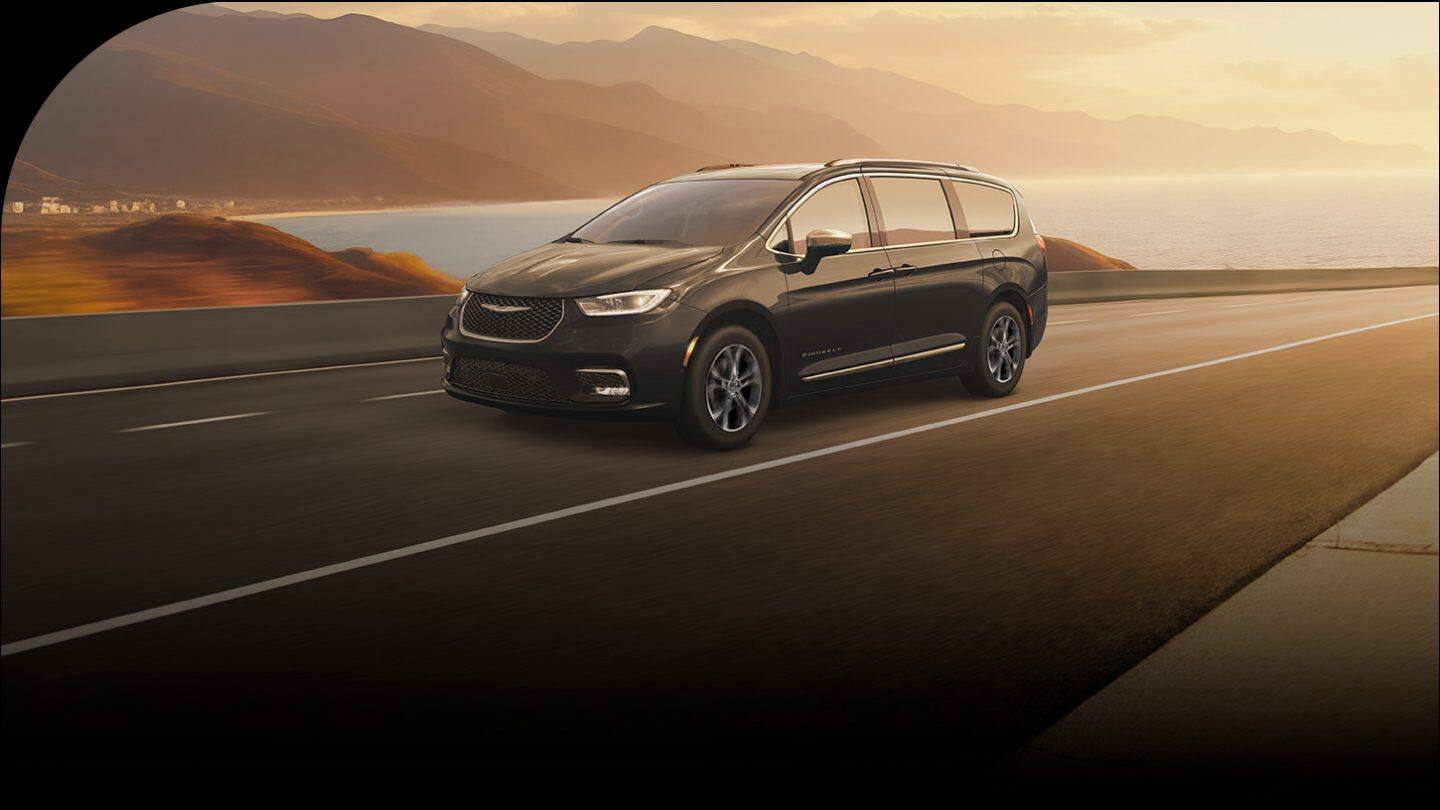 A black 2026 Chrysler Pacifica Pinnacle traveling down a highway at sunset, with a lake and mountains in the background.