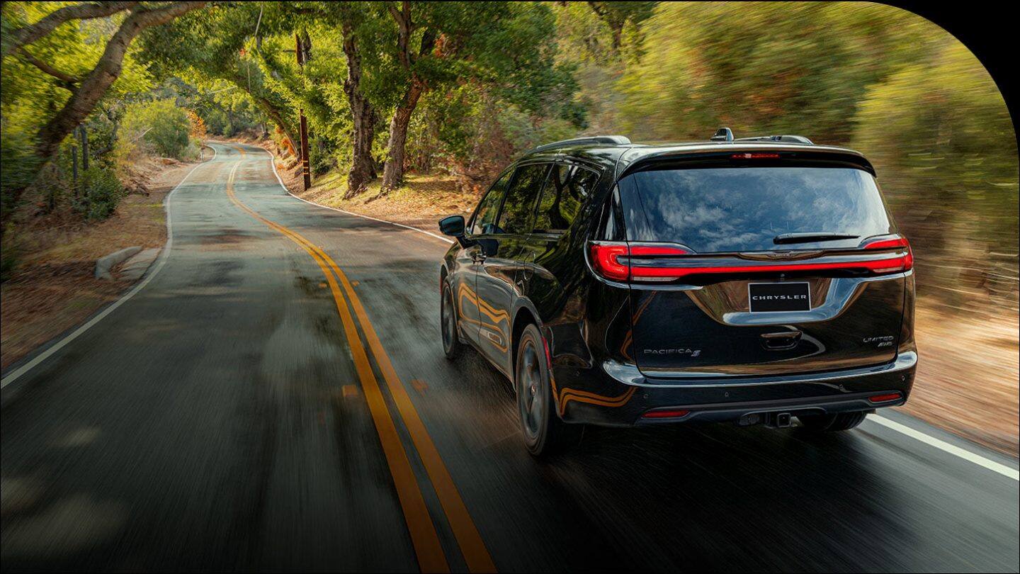 A rear angle of a black 2026 Chrysler Pacifica Limited AWD traveling down a country road. The background is blurred to indicate the vehicle is in motion.