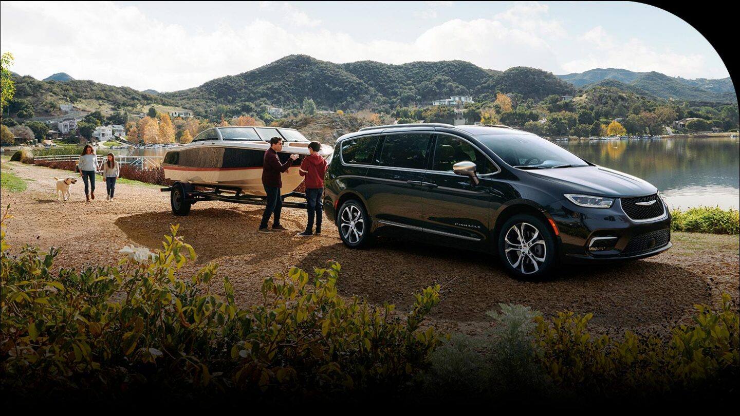 A black 2026 Chrysler Pacifica Pinnacle parked beside a lake, towing a motorboat, with a family of four nearby.