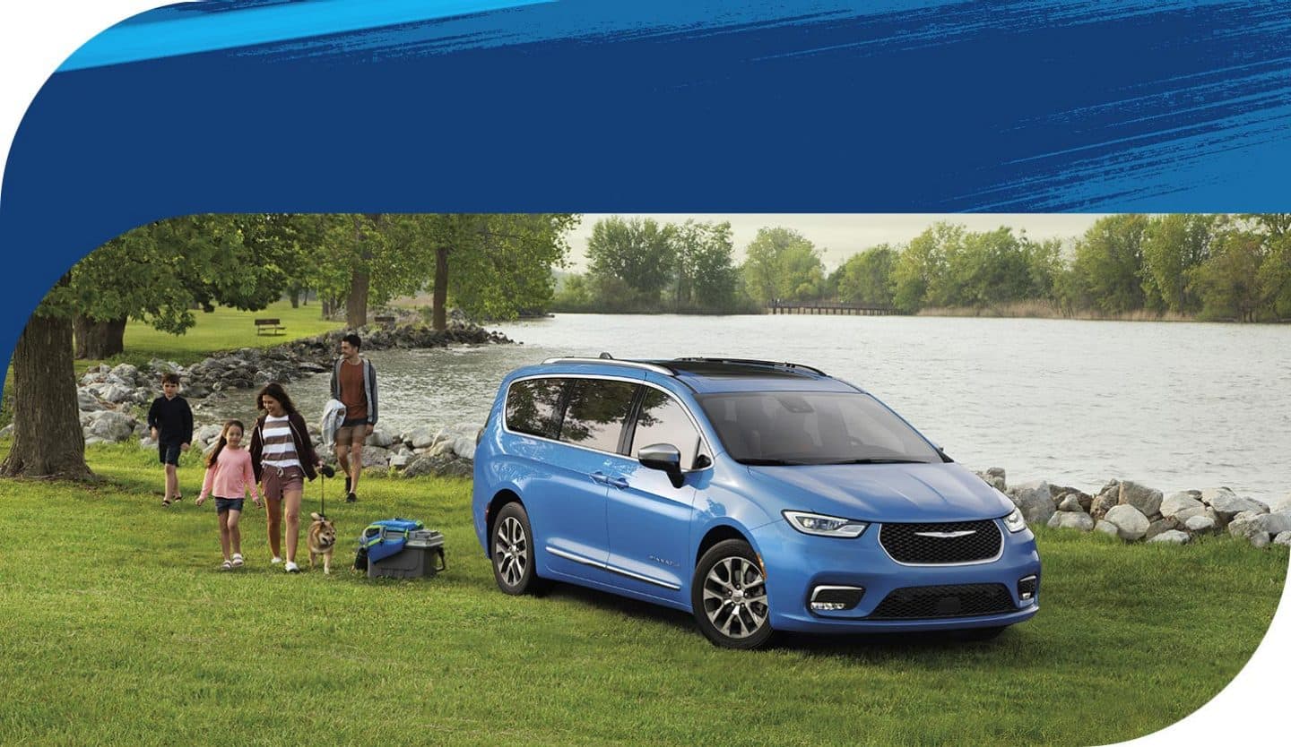 A blue 2026 Chrysler Pacifica Plug-in Hybrid Pinnacle parked beside a lake with a family of four walking nearby.