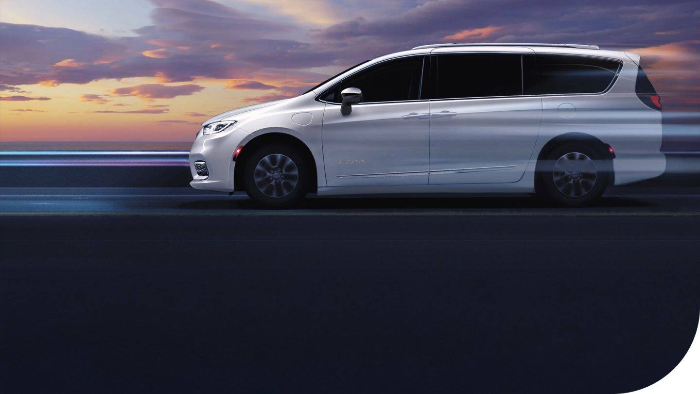 A driver-side profile of a white 2026 Chrysler Pacifica Plug-in Hybrid Pinnacle with its wheels blurred to indicate the vehicle is in motion.