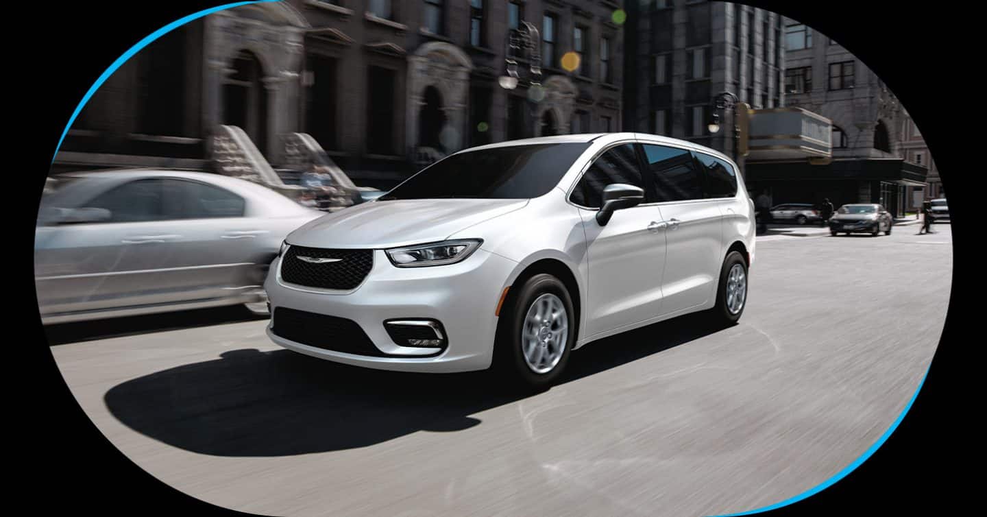 A white 2026 Chrysler Pacifica Select traveling down a busy city street.