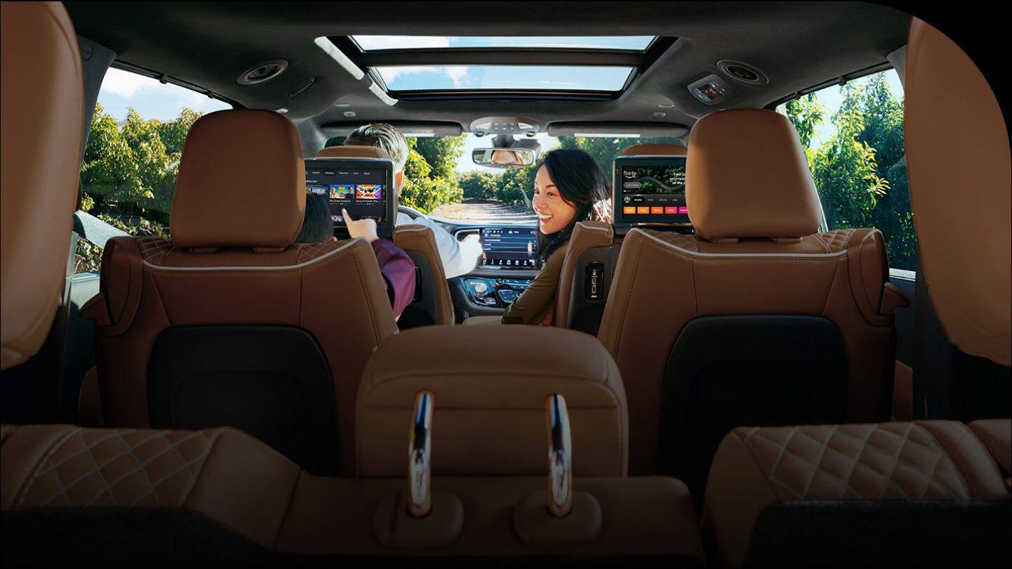 The interior of the 2026 Chrysler Pacifica Pinnacle from behind the third-row perspective forward, focusing on the front passenger turning to look at a child in the second row who is browsing options on the front seatback-mounted touchscreen.