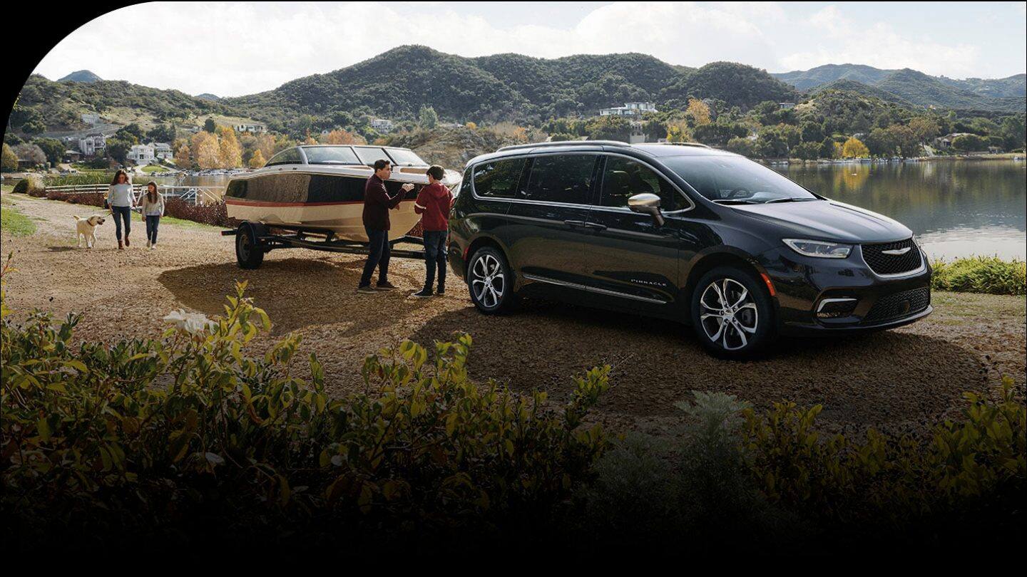 A black 2026 Chrysler Pacifica Pinnacle parked beside a lake, towing a motorboat, with a family of four nearby.