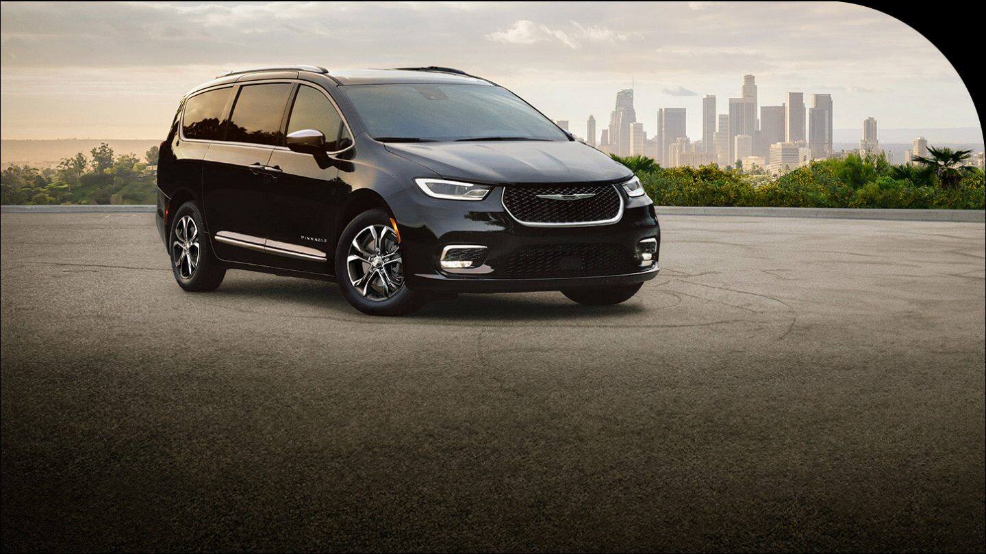 A passenger-side front angle of a black 2026 Chrysler Pacifica Pinnacle parked on a lot with a metropolitan skyline in the background.