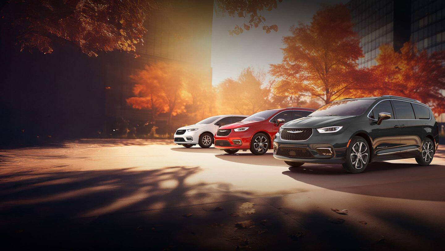 Three 2026 Chrysler minivan models parked on an oceanside beach. From left to right: a white Chrysler Voyager, a blue Pacifica Pinnacle Plug-in Hybrid and a red Pacifica Pinnacle. Chrysler Pacifica sales event.