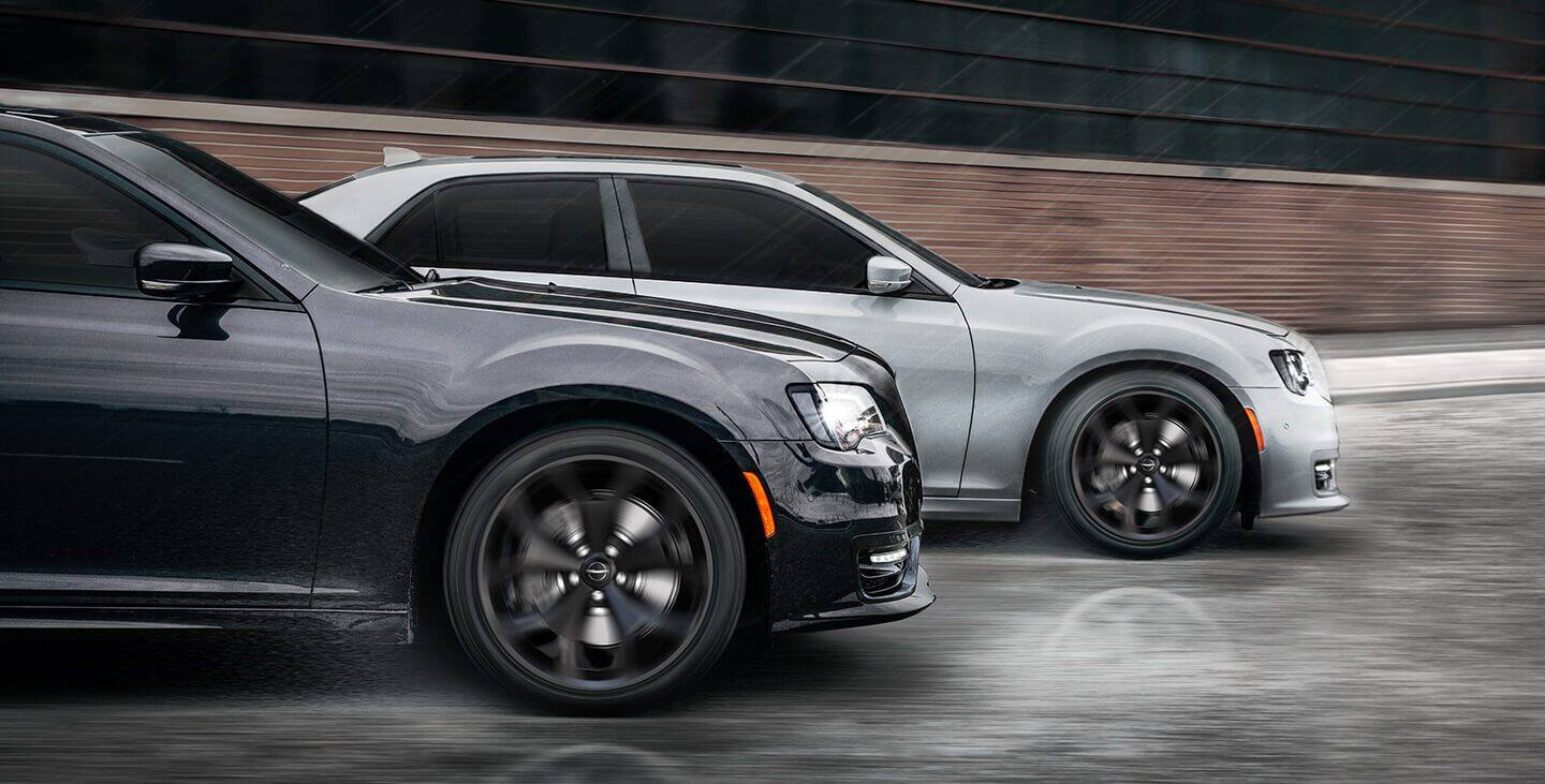 Display Two 2021 Chrysler 300S V8 models, one in Granite Crystal—a glossy gray, and the other in Silver Mist, being driven on a city street in the rain.