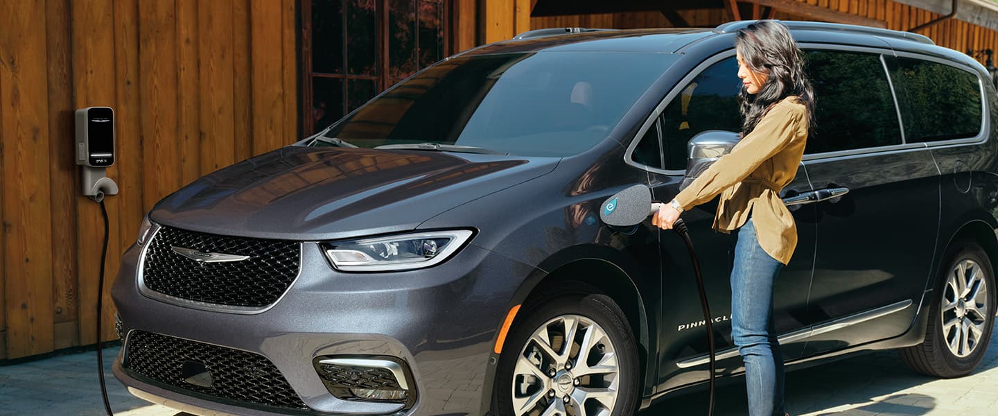 A woman beside a 2022 Chrysler Pacifica Pinnacle Hybrid plugging a charging cable into the vehicle's charging port.