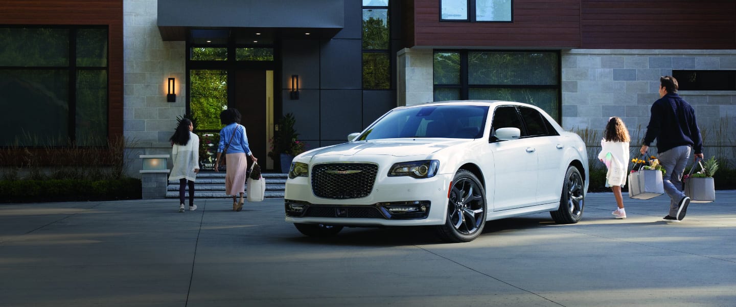 A 2023 Chrysler 300 parked in the driveway of a large, modern house with a family of four walking from the vehicle to the front door.
