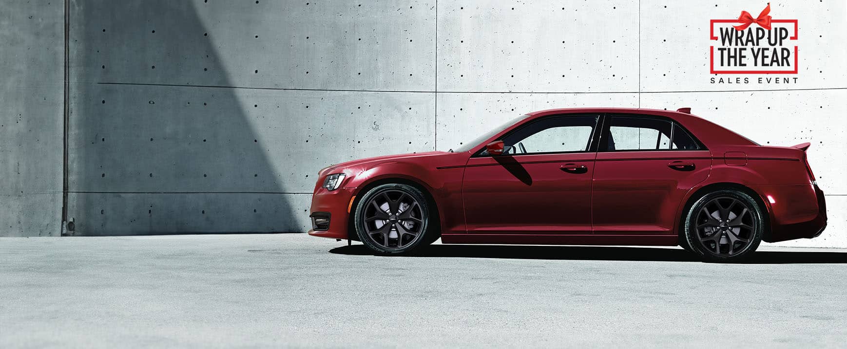 A driver-side profile of a red 2023 Chrysler 300S V8 parked next to a cement wall. Wrap up the year Sales Event logo.