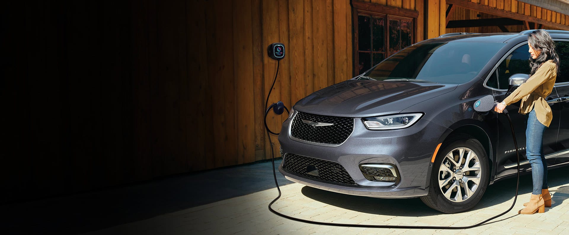 A rear view of the 2023 Chrysler Pacifica TBD Hybrid parked outside a garage, with a charging cord plugged into its charging port.