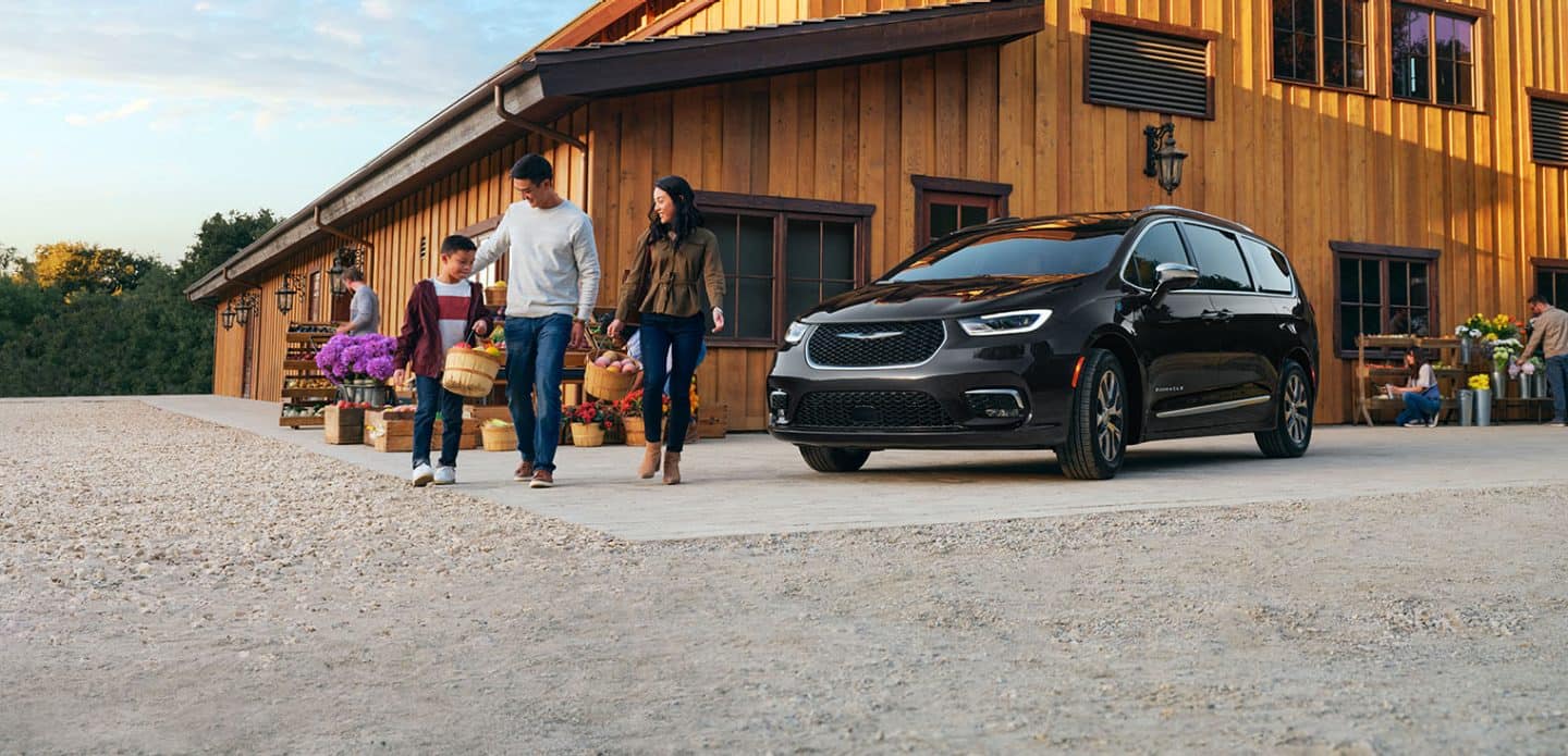 Display A black 2023 Chrysler Pacifica TBD Hybrid parked beside a farmers market with a family of three walking nearby, carrying bushels of produce.