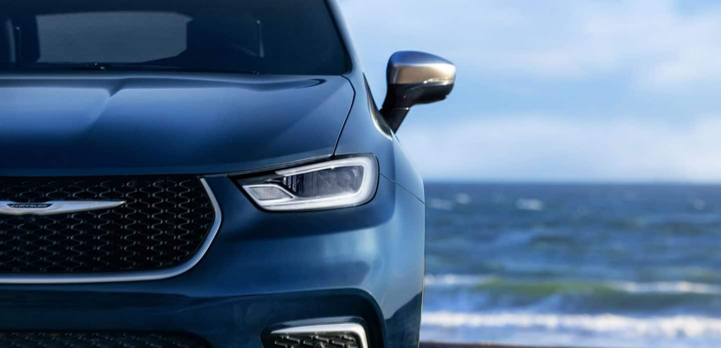 Display A close-up of the grille and headlamp on a blue 2023 Chrysler Pacifica Pinnacle Hybrid parked beside a body of water.