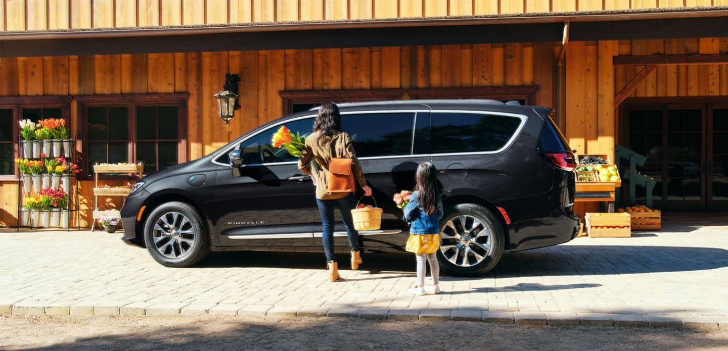 Display A mother and daughter with their hands full standing beside the hands-free power sliding side door on a black 2023 Chrysler Pacifica Pinnacle Hybrid parked at a farmers market, as the mother swings her foot under the door.
