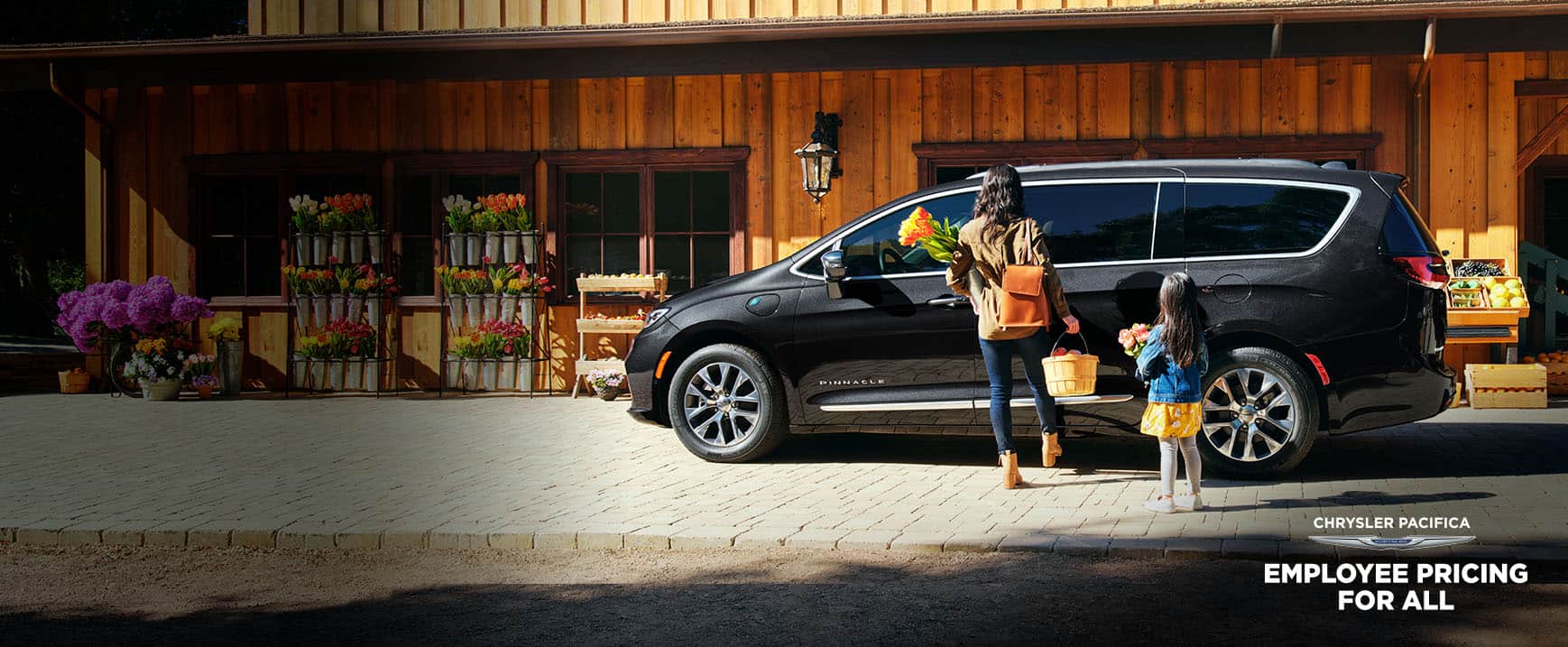 A mother and daughter with their hands full standing beside the hands-free power sliding side door on a black 2023 Chrysler Pacifica Pinnacle Hybrid parked at a farmers market, as the mother swings her foot under the door. Employee Pricing for All logo.
