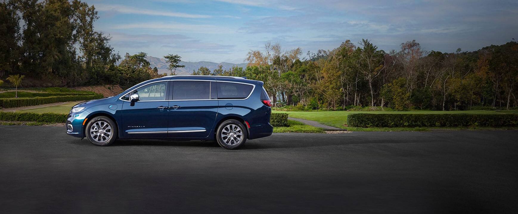The driver-side profile of a 2023 Chrysler Pacifica Pinnacle Plug-in Hybrid parked in a lot beside a park with mountains in the background. 