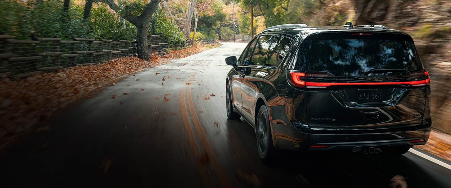 The rear view of a 2023 Chrysler Pacifica Limited with S Appearance Package being driven down a tree-lined curved country road in autumn. The background is blurred to indicate the speed of the vehicle. 