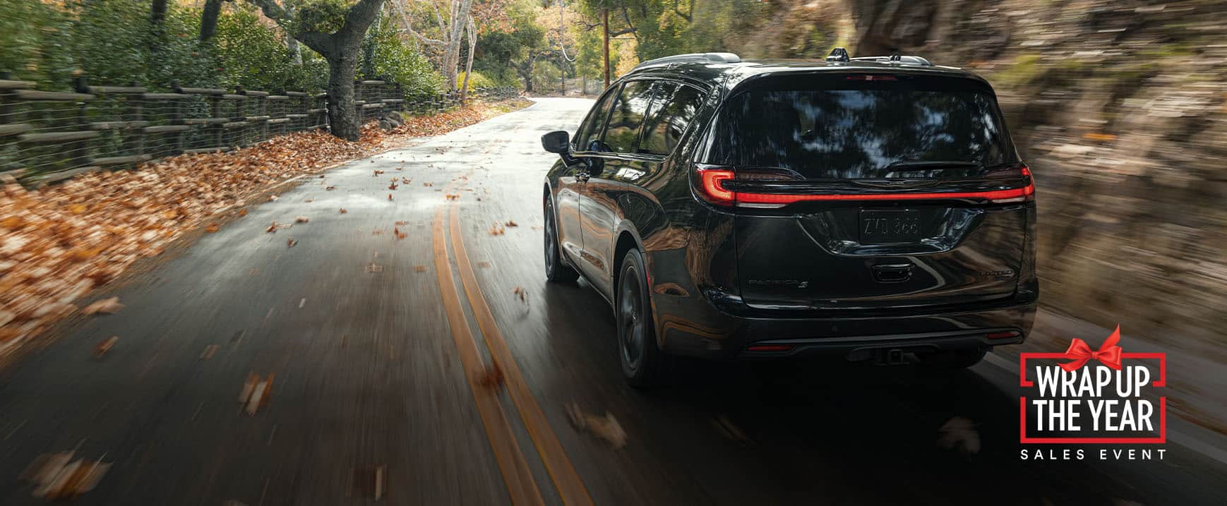 A rear angle of a 2023 Chrysler Pacifica Limited with the S Appearance Package being driven on a country road. The background is blurry indicating the vehicle is in motion. Wrap up the year Sales Event logo.