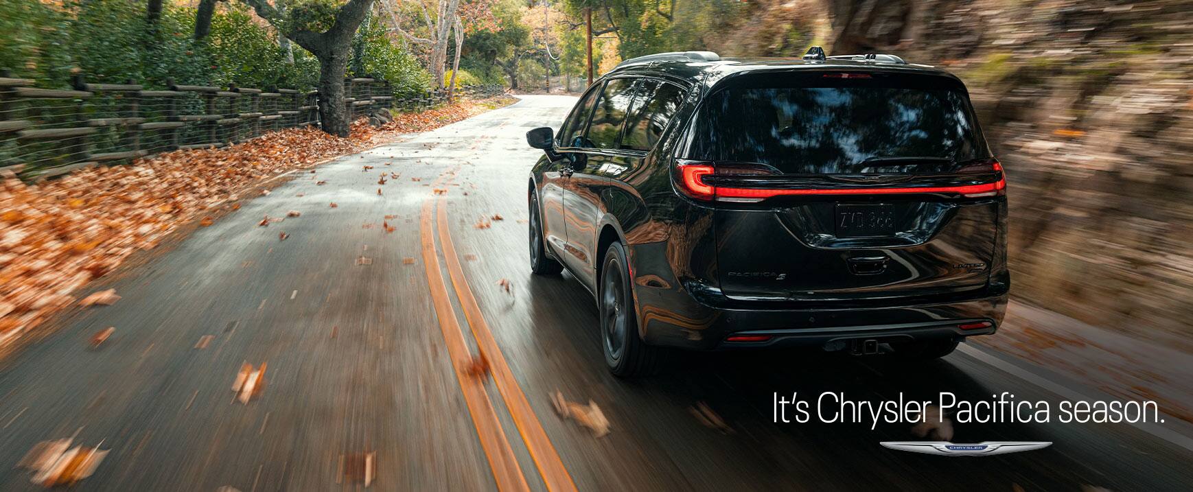 A rear angle of a 2023 Chrysler Pacifica Limited with the S Appearance Package being driven on a country road. The background is blurry indicating the vehicle is in motion. It's Chrysler Pacifica Season logo. 