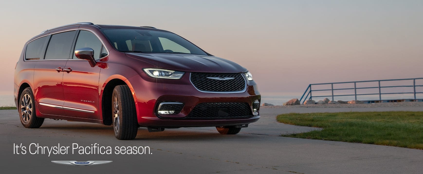 An angled passenger-side profile of a red 2023 Chrysler Pacifica Pinnacle Plug-In Hybrid parked in a waterfront lot. It's Chrysler Pacifica Season logo. 