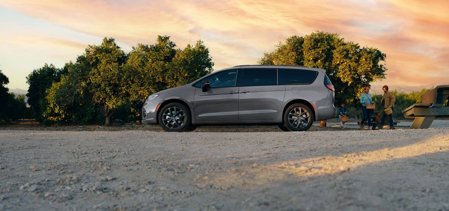 Display A driver-side profile of a silver 2024 Chrysler Pacifica Limited with S appearance Package parked beside an orchard.