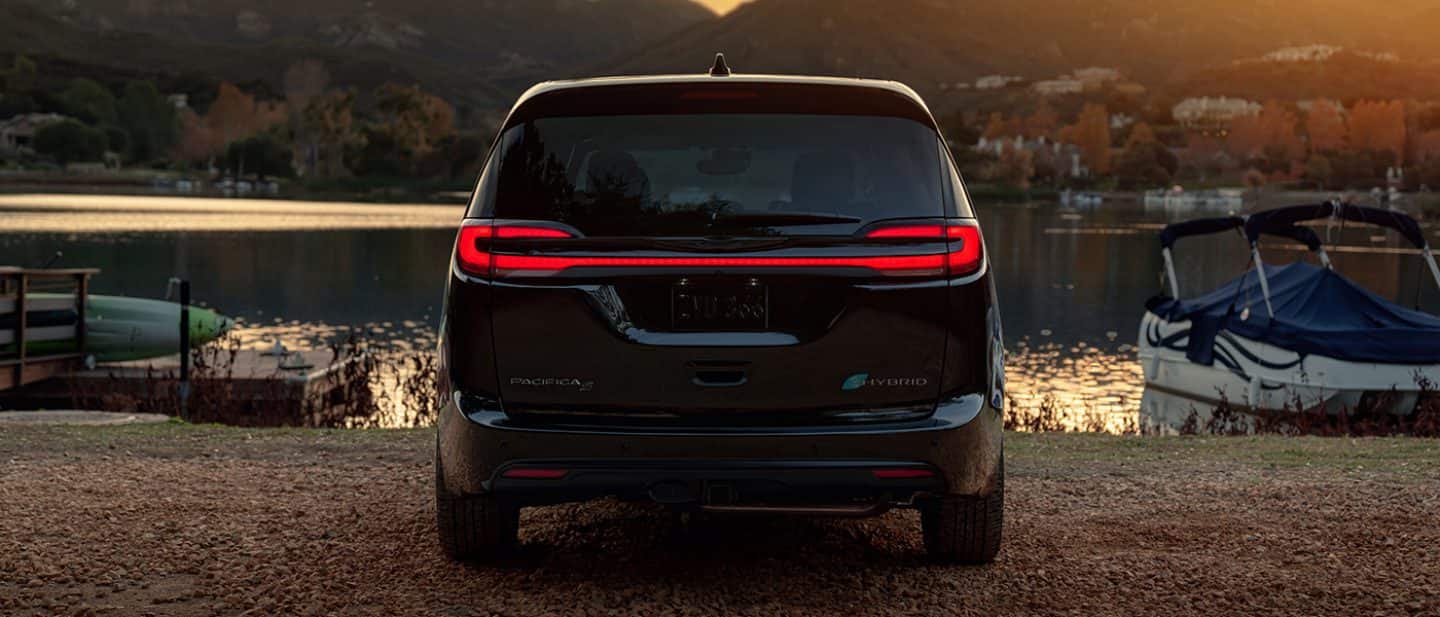 Display The rear of the 2024 Chrysler Pacifica Limited with S Apearance Package Plug-In Hybrid with its full-width tailllamps lit, parked beside a lakeside dock at dusk.