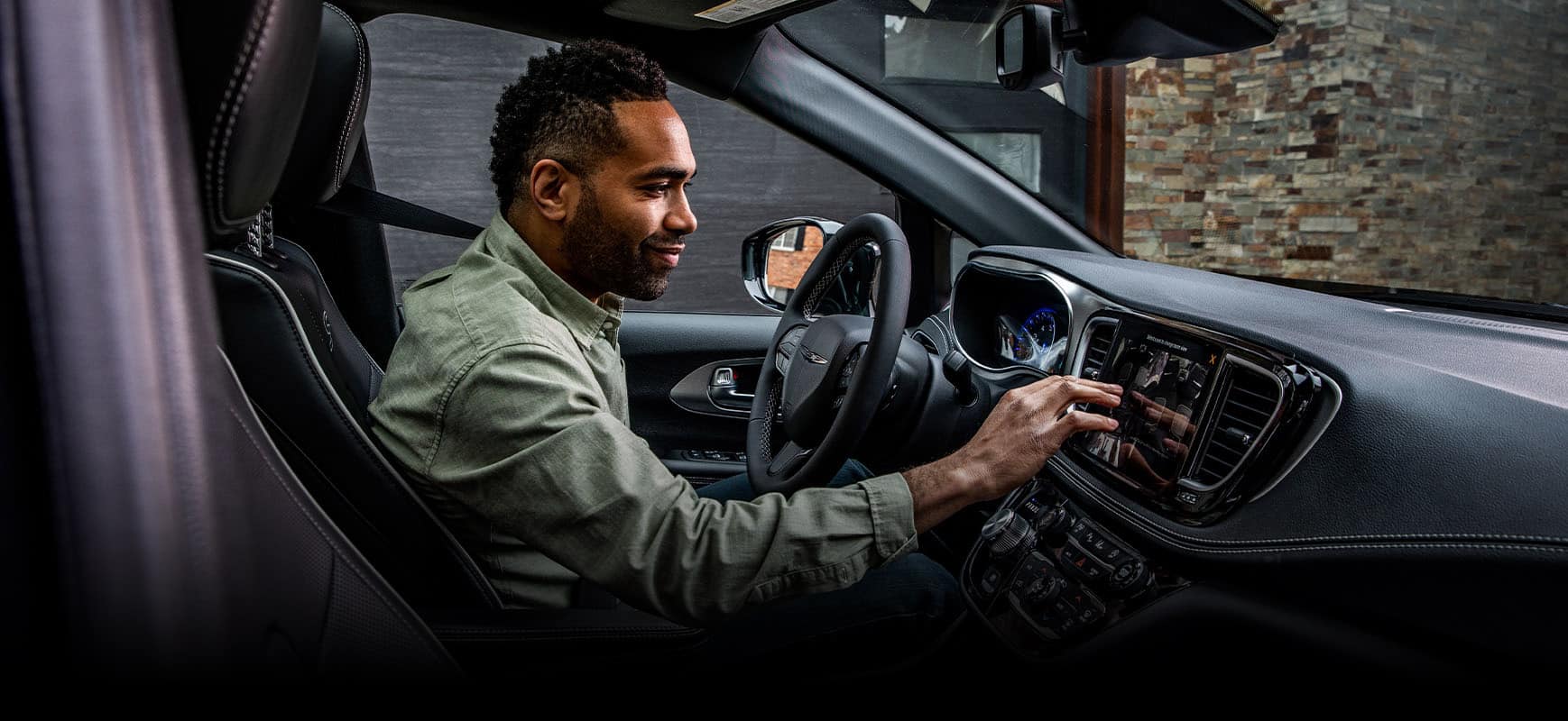 The front seats in the 2024 Chrysler Pacifica Plug-In Hybrid with the driver looking at the Uconnect touchscreen as he makes a selection.