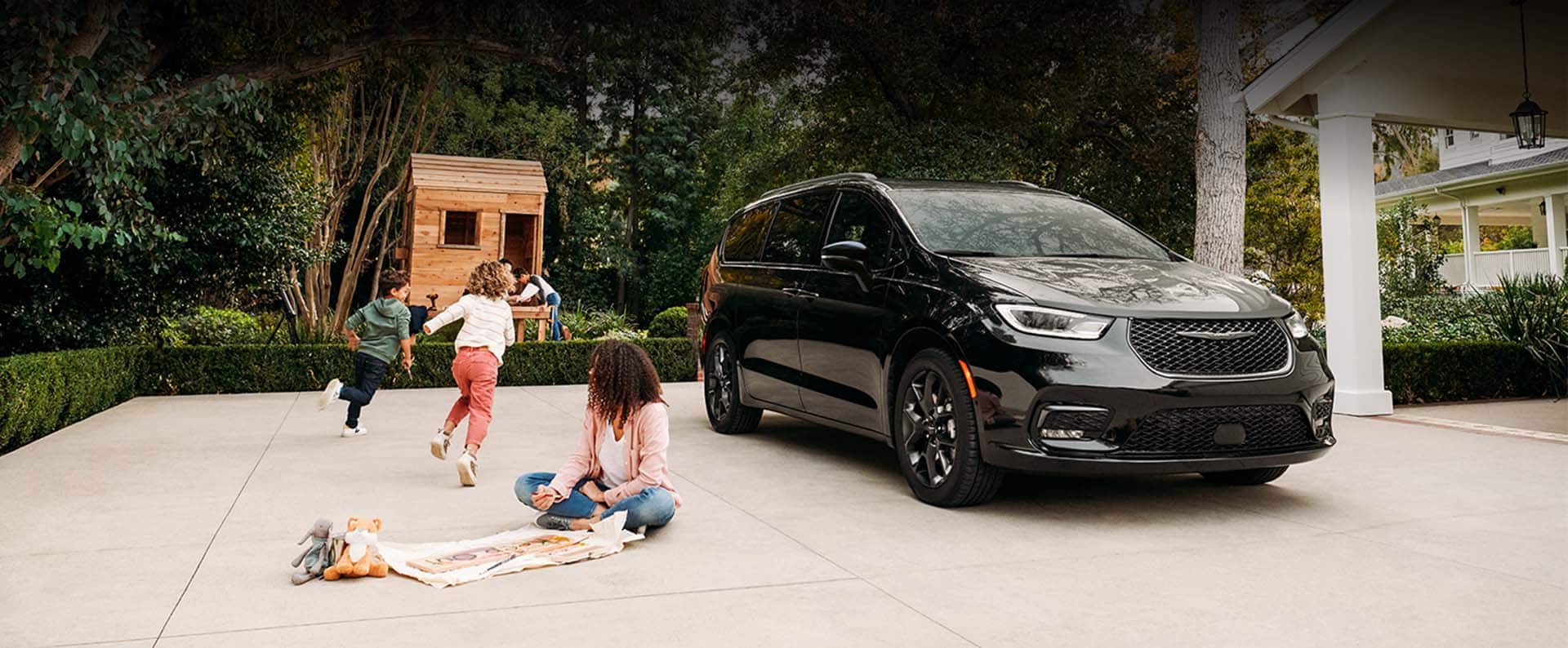 A black 2024 Chrysler Pacifica Limited with S Appearance Package parked in the driveway of a residential home with children playing beside it.