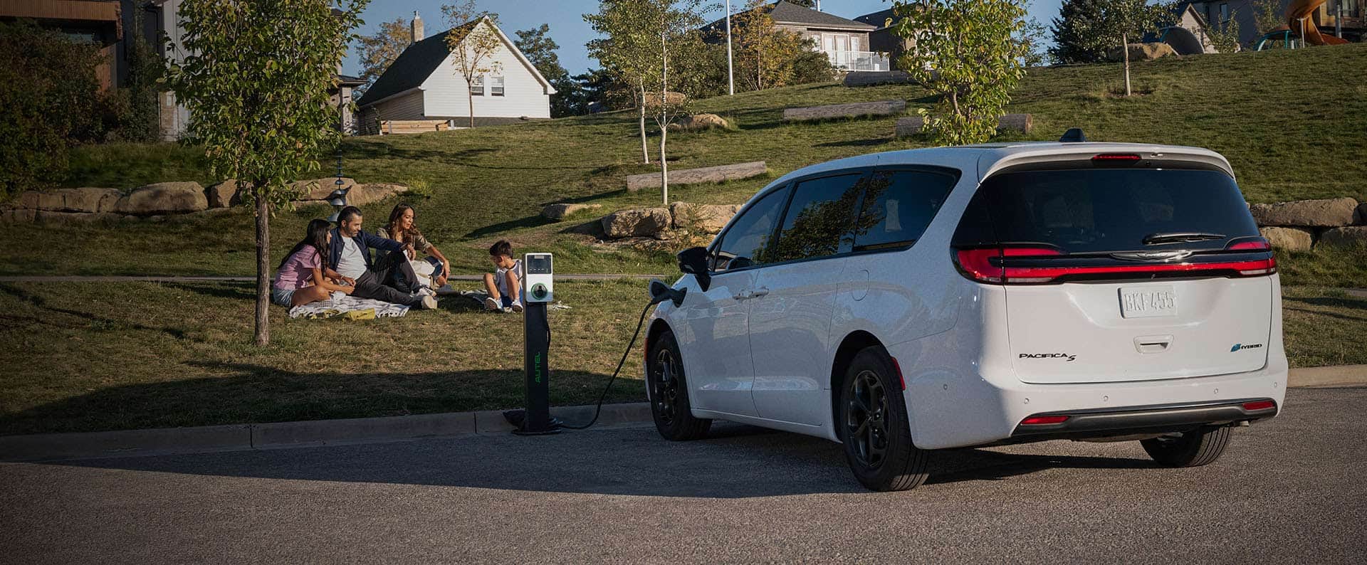 An angled rear driver-side profile of a white 2024 Chrysler Pacifica Plug-in Hybrid S Appearance parked beside a PHEV charging station with the charging cord plugged into the vehicle’s charging port, while a family of four sit on the grass nearby.