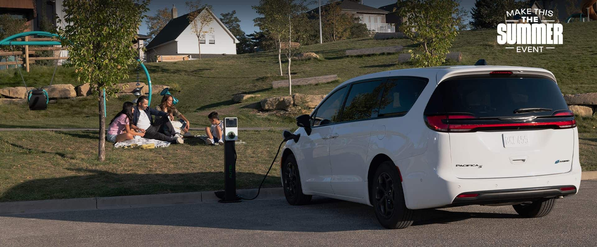 An angled rear driver-side profile of a white 2024 Chrysler Pacifica Plug-in Hybrid S Appearance parked beside a PHEV charging station with the charging cord plugged into the vehicle’s charging port, while a family of four sit on the grass nearby.