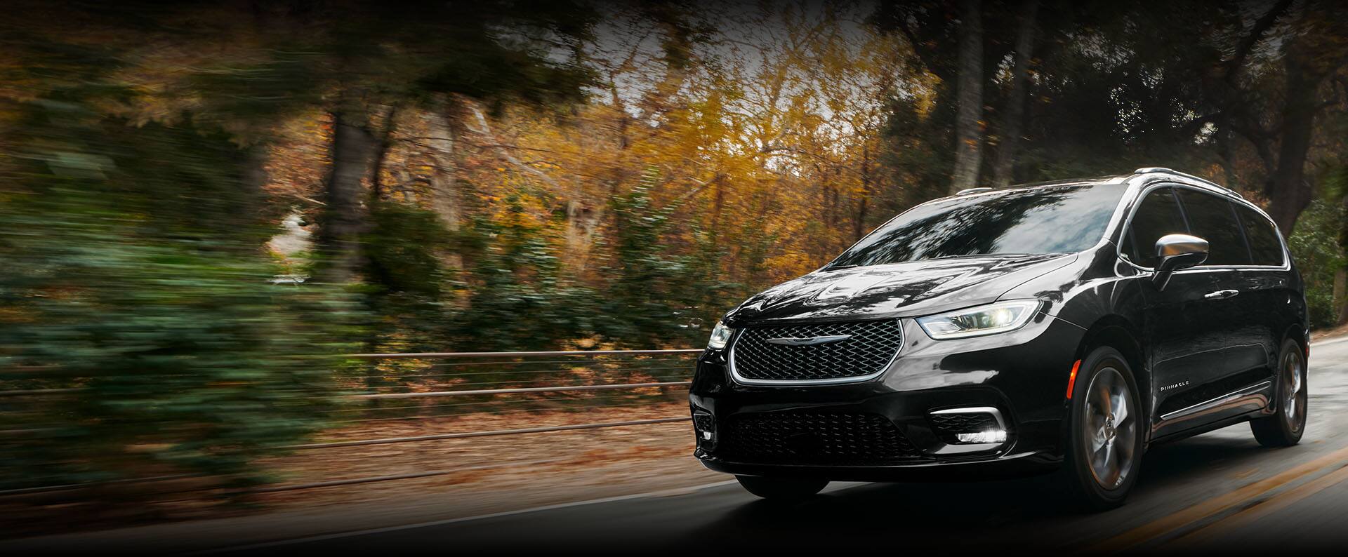 An angled driver-side profile of the 2024 Chrysler Pacifica Pinnacle being driven down a country road, the background is blurred to indicate the vehicle is in motion.