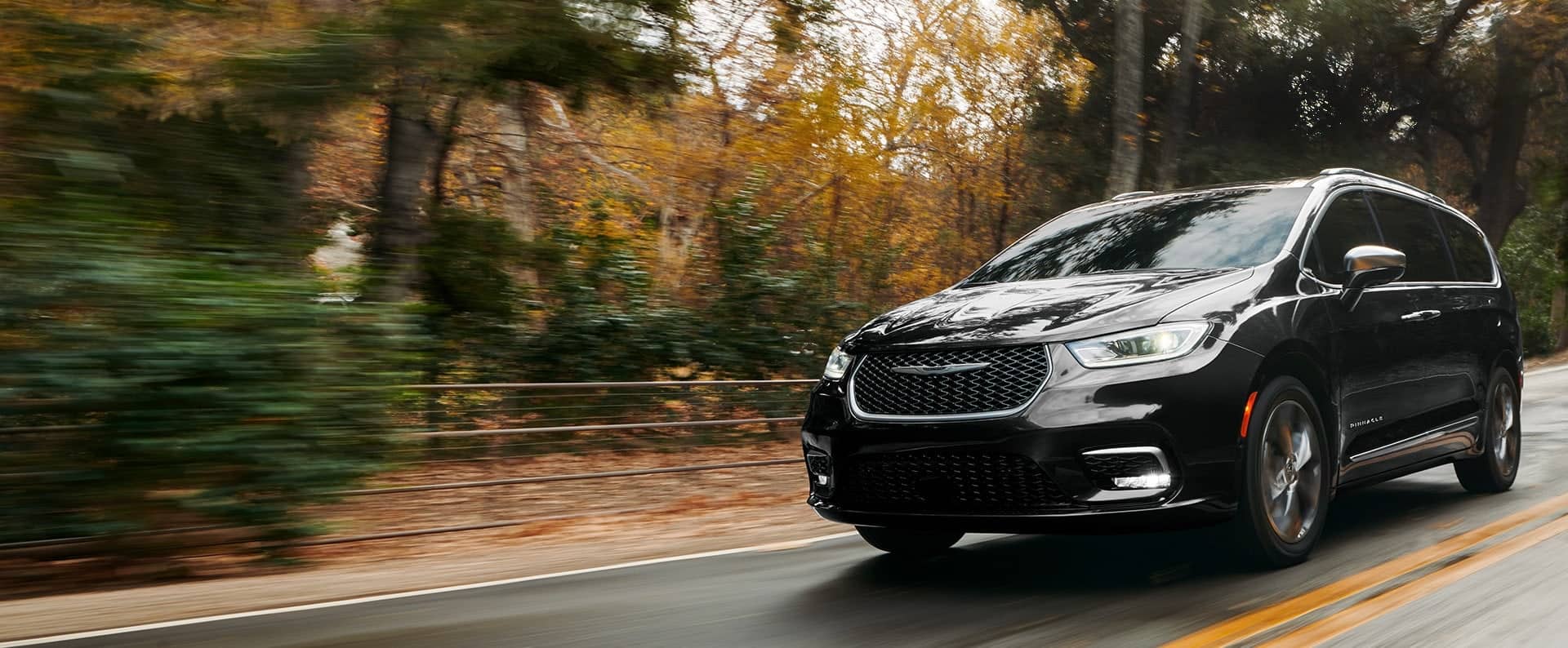 An angled driver-side profile of the 2024 Chrysler Pacifica Pinnacle being driven down a country road, the background is blurred to indicate the vehicle is in motion.