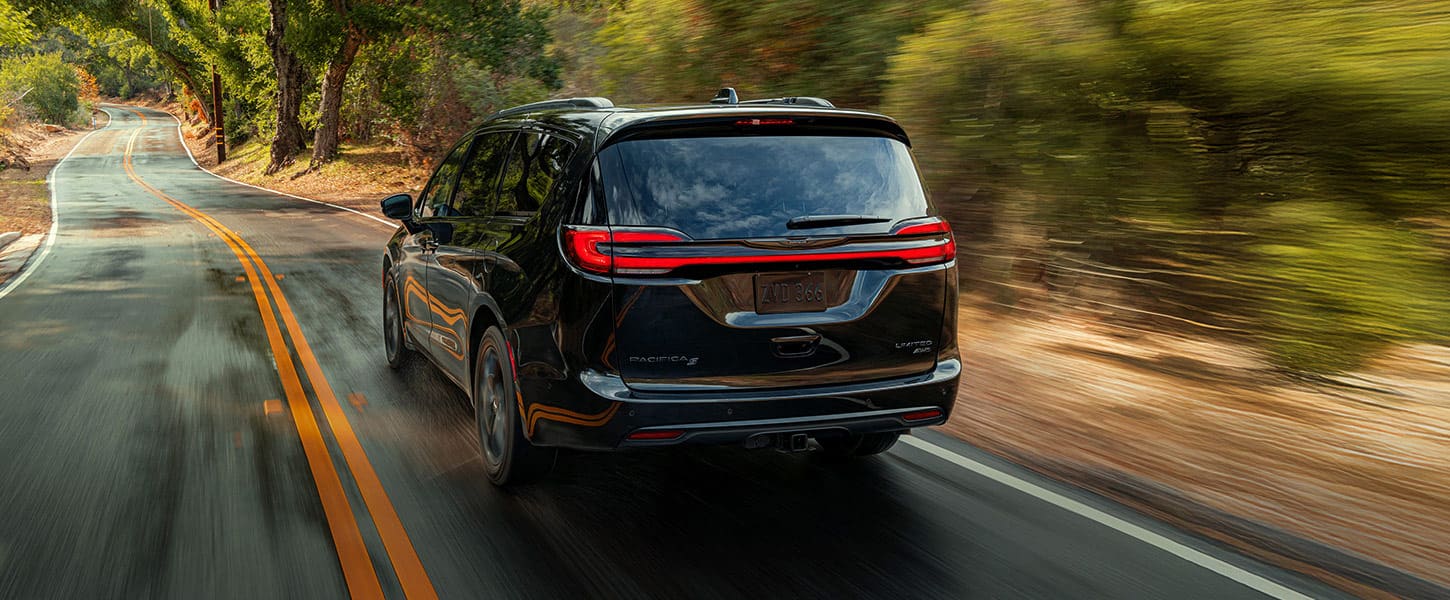 An rear driver-side profile of the 2025 Chrysler Pacifica Pinnacle being driven down a country road, the background is blurred to indicate the vehicle is in motion.