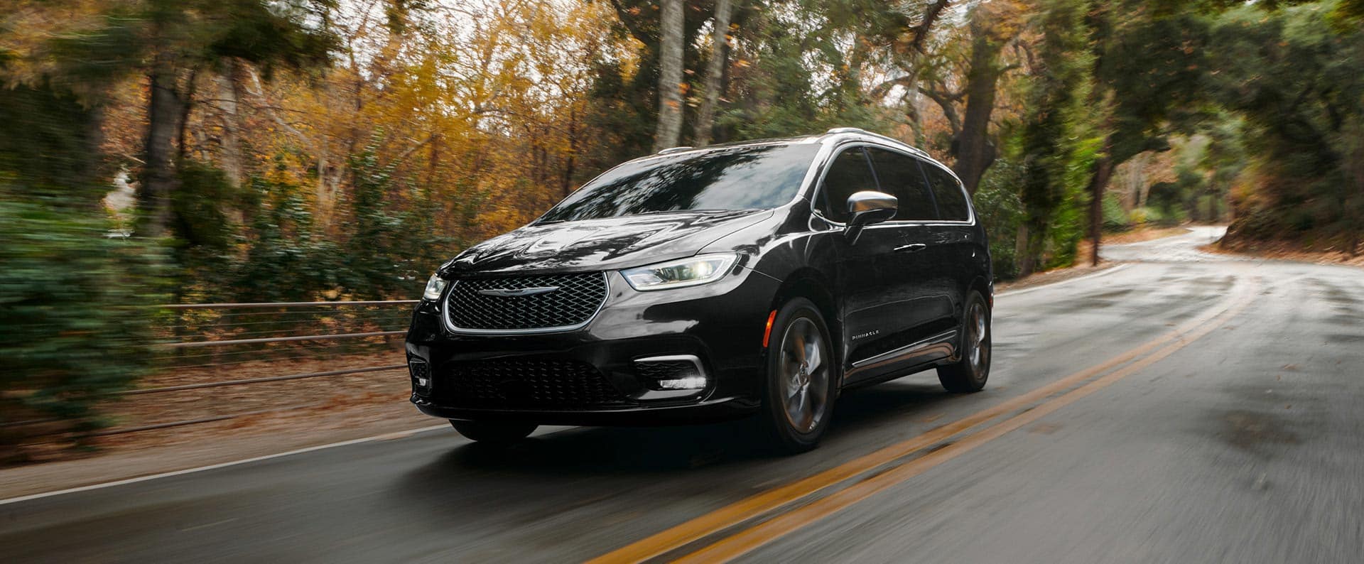 An angled driver-side profile of the 2025 Chrysler Pacifica Pinnacle being driven down a country road, the background is blurred to indicate the vehicle is in motion.
