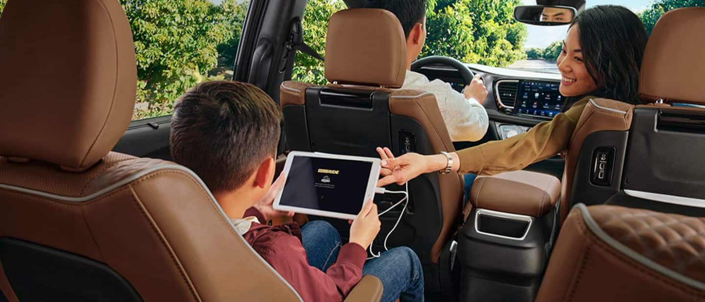 Display The interior of the 2025 Chrysler Pacifica Pinnacle with a man driving, a woman reaching into the rear and a boy in the second row holding a tablet that is plugged into an outlet.
