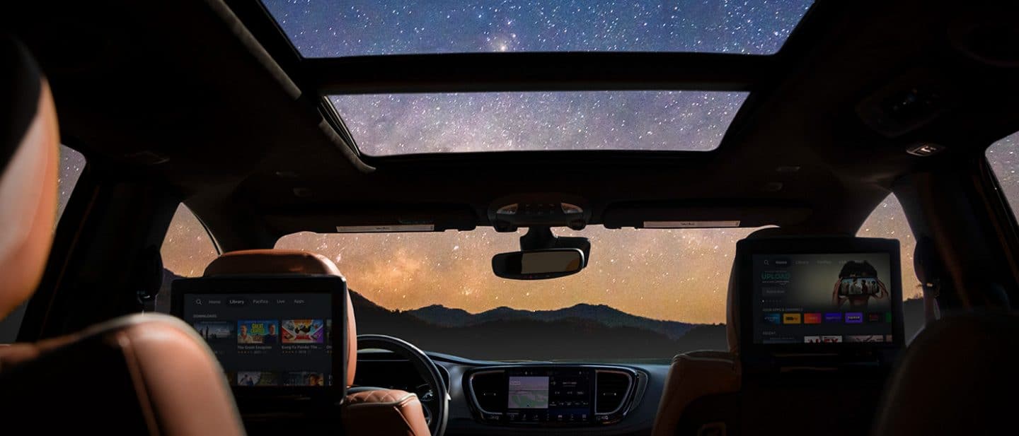 Display The dual-pane sunroof in the 2025 Chrysler Pacifica Pinnacle, open revealing the night sky loaded with stars, as the two seatback-mounted touchscreens display a variety of selections.