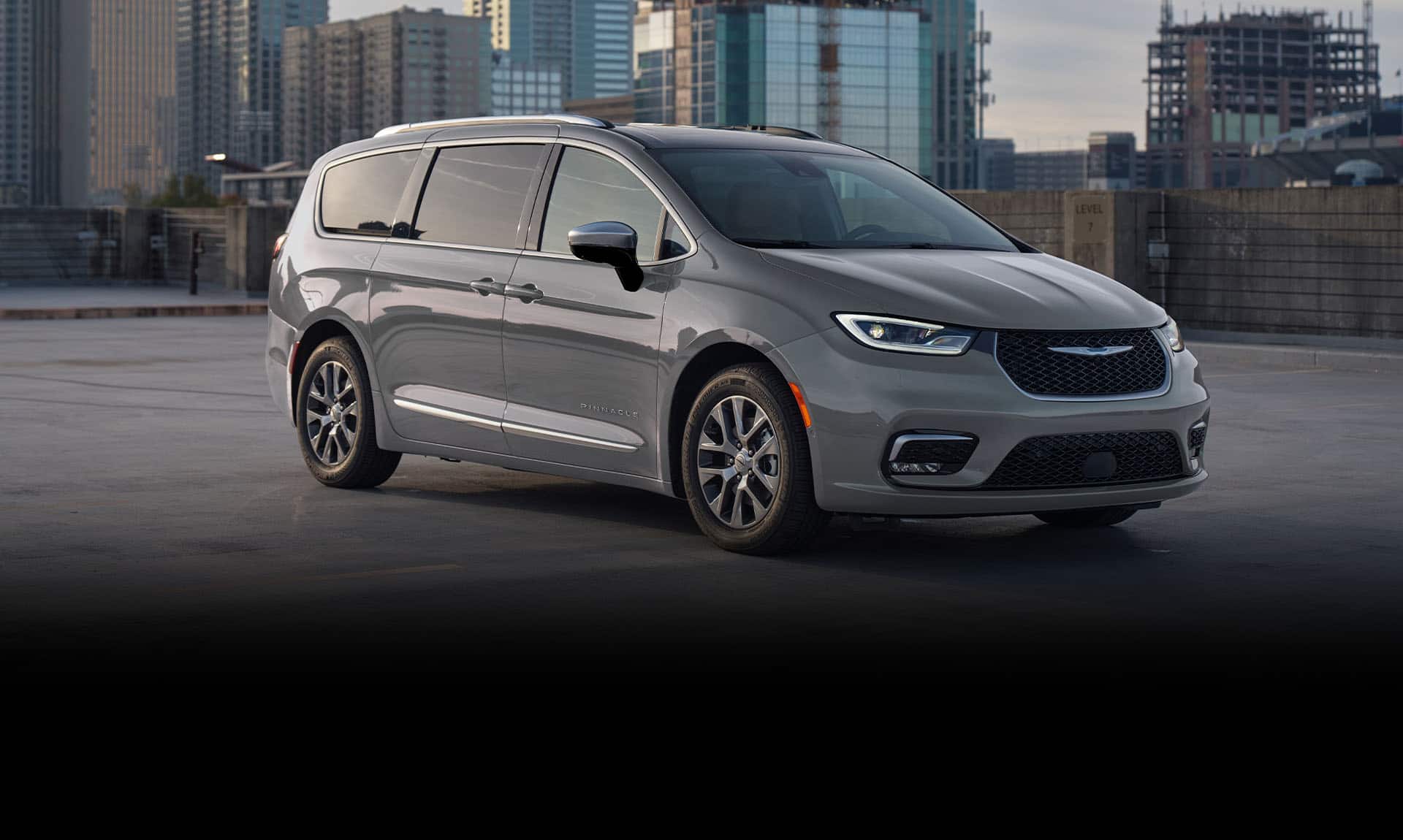 A passenger-side front angle of a gray 2025 Chrysler Pacifica Pinnacle Plug-in Hybrid parked on the roof of a parking structure with tall buildings in the background.