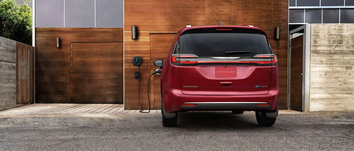 Display A rear angle of a red 2025 Chrysler Pacifica Pinnacle Plug-in Hybrid parked outside a garage, with a charging cord plugged into its charging port.